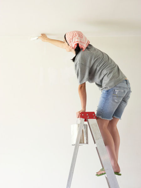 Young woman standing on step ladder, painting wall, side view